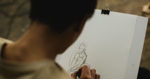Artist making a loose charcoal sketch of a seated figure on paper clipped to a drawing board