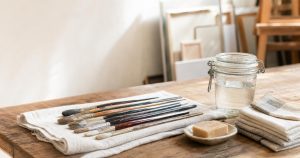 Freshly washed oil paint brushes drying flat on a linen cloth beside a lidded jar and a bar of brush soap on a studio table.
