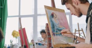 Painter working on a seated figure study at an easel in a bright studio, with other students in the background
